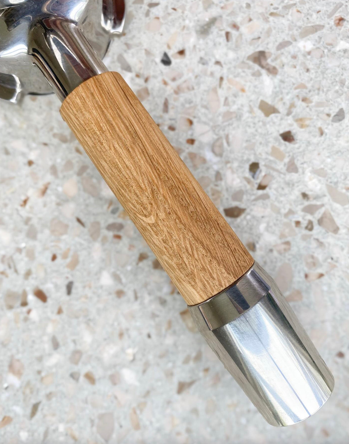 Detaljbild på en rostfri portafilter med handtag i ljus trä mot en slät, mönstrad bänkskiva.